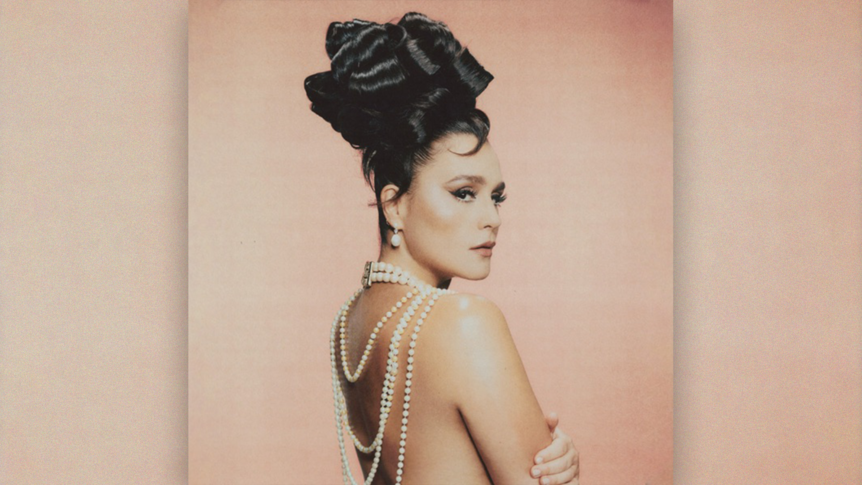 Topless Jessie Ware with her back to camera and a string of pearls falling down her back. Her black hair is done up, arms folded