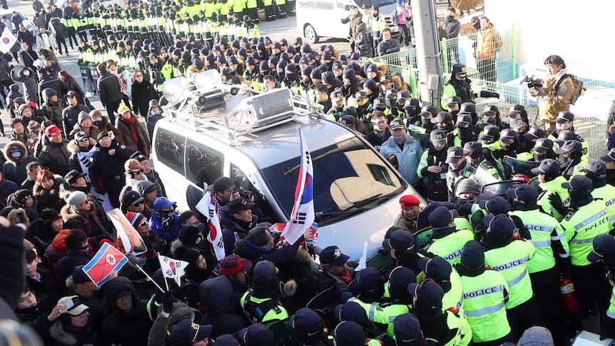 A crowd of protestors and another crowd of police surround a white van.