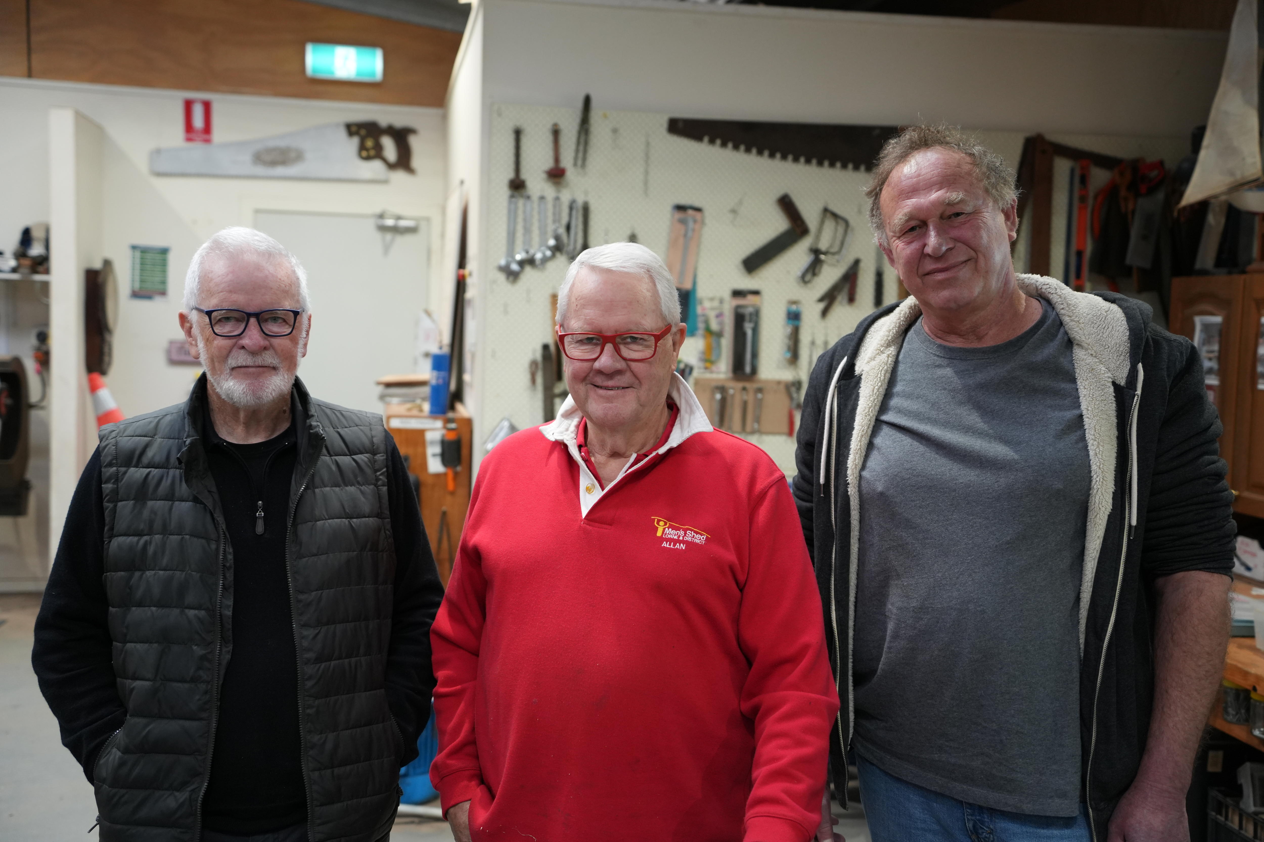 Two white haired men with glasses stand beside a man with curly hair inside a shed with tools.