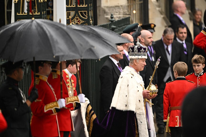 King Charles holding an orb and scepter wearing a crown. He is surrounded by guards holding umbrellas. 