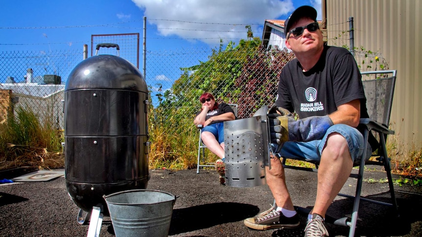 A man and woman sit outdoors by a barbecue smoker