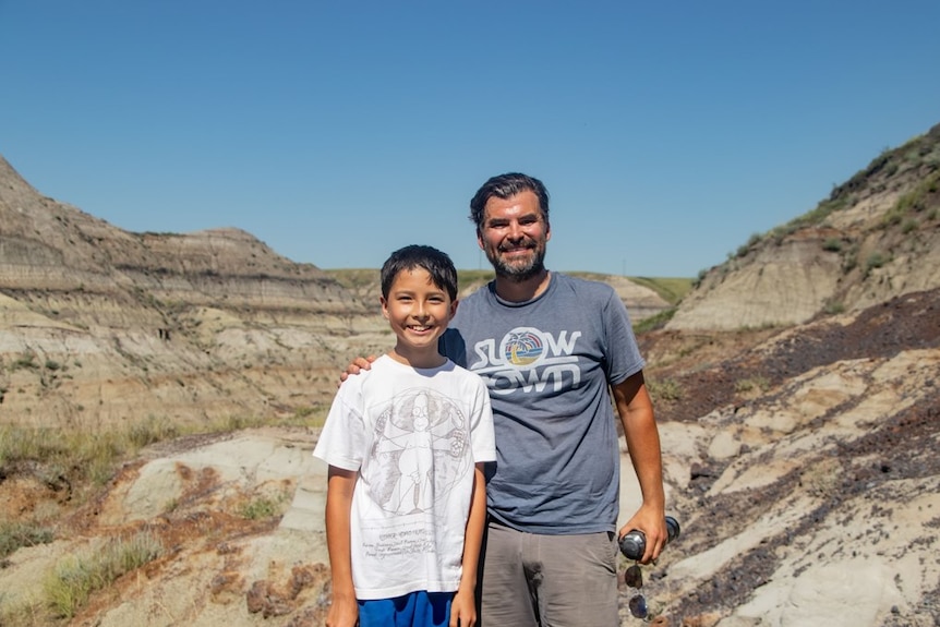 A boy and a man smile as they pose for a photograph at the head of a deep and long desert canyon.