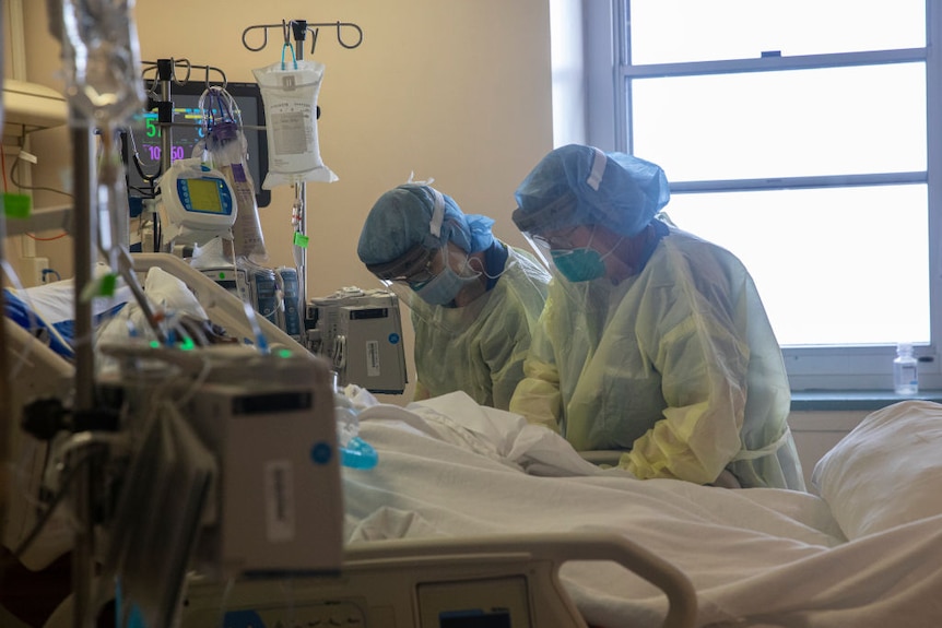 Two people wearing medical capes, caps and face shields work on a person on a hospital bed with drips and machines connected.