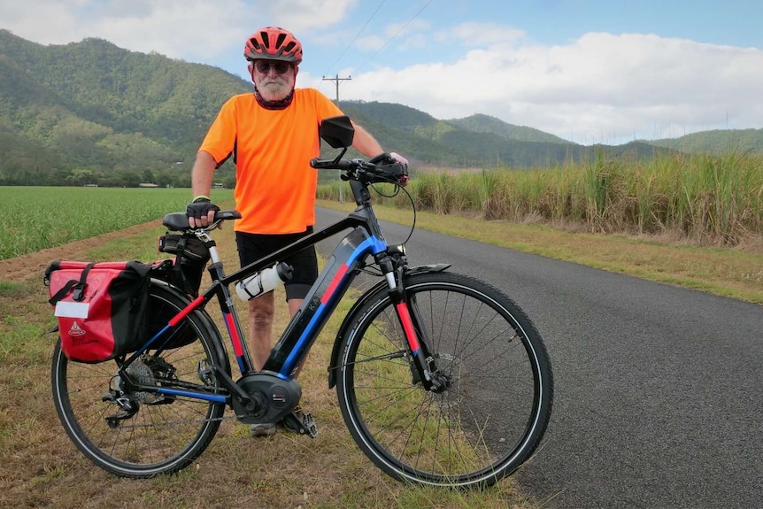 Cyclist with white beard standing behind a bicycle with sugar cane fields in the background