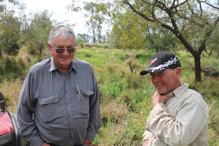 Two men are pictured standing next to each other. Behind them bushy land, with a tree on the right.