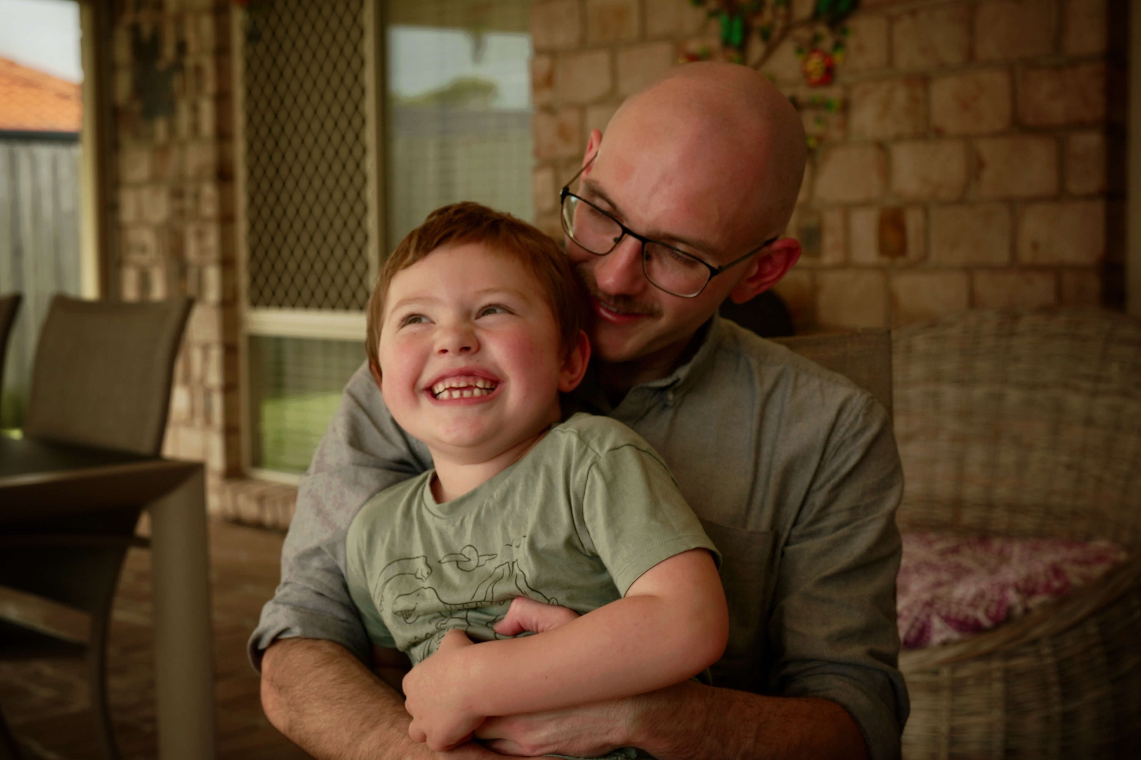 A young boy smiles while sitting in his dad's lap who looks down at him.