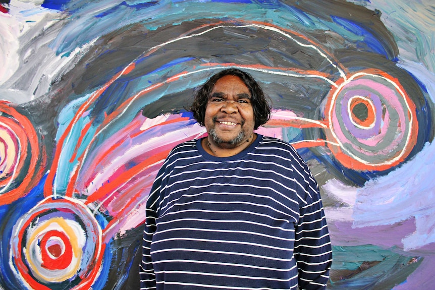 Colour photograph of artist Barbara Mbitjana Moore standing in front of one her vibrant paintings.