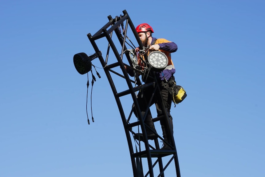 A man on a ladder holding a light and trying to install it outdoors in a park