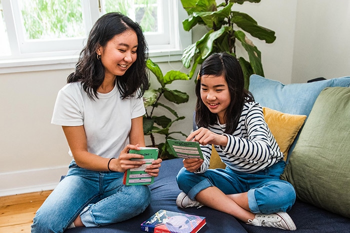 A woman helps a young girl play a card game while sitting on a couch in a loungeroom.