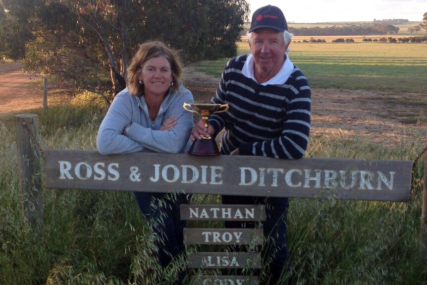 A woman and man lean on a farm sign with a trophy.