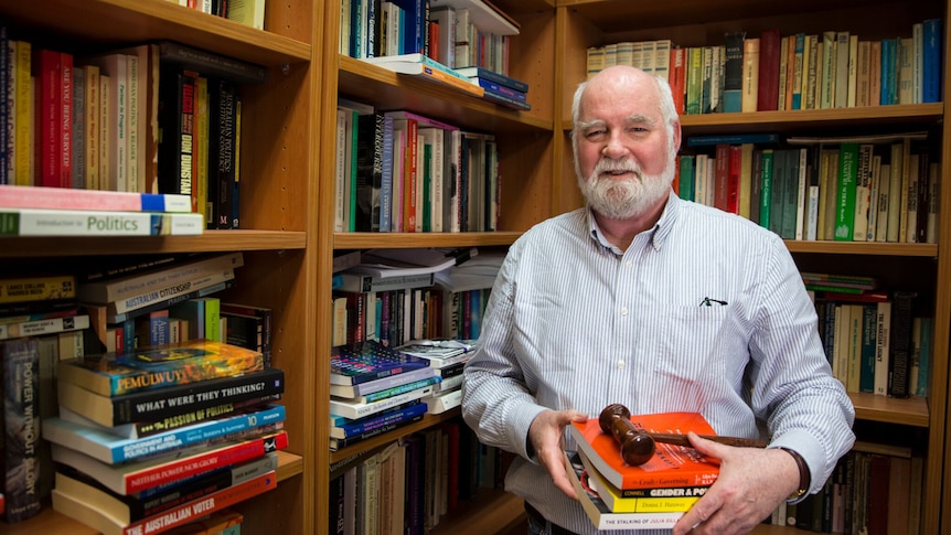A man with a white beard holds a stack of books in front of a bookcase.