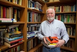 A man with a white beard holds a stack of books in front of a bookcase.
