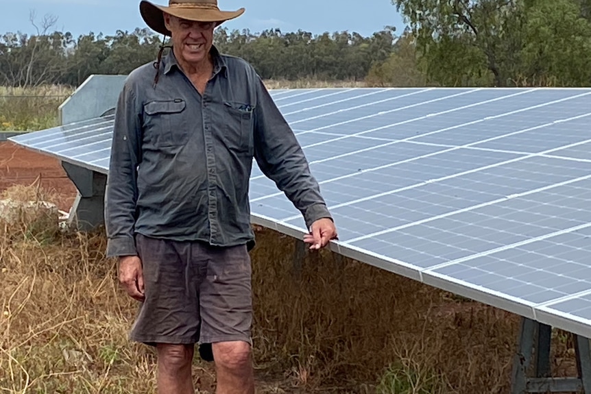 A man wearing a navy shirt and wide brimmed hat stands beside a large solar panel in a paddock. 