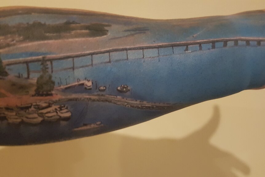 A tattoo on a man's forearm depicting a bridge over a blue river, with boats on the side.
