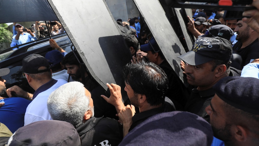 A large crowd with people holding shields over some people exiting a car. 