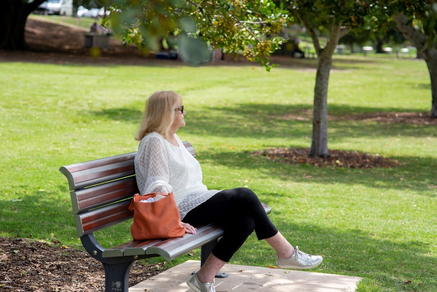 A woman with blonde hair and dark sunglasses sits on a park bench on a sunny day, looking off to her left into the distance.