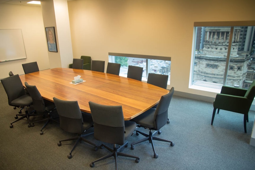 A white room with an oblong table and 12 chairs, plus two armchairs. Box of tissues on table. Whiteboard in background.