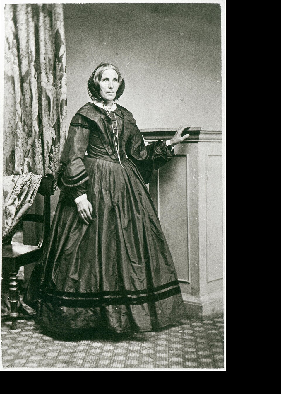 balck and white historic image of a woman in long dress posing against a wall with curtains to the left