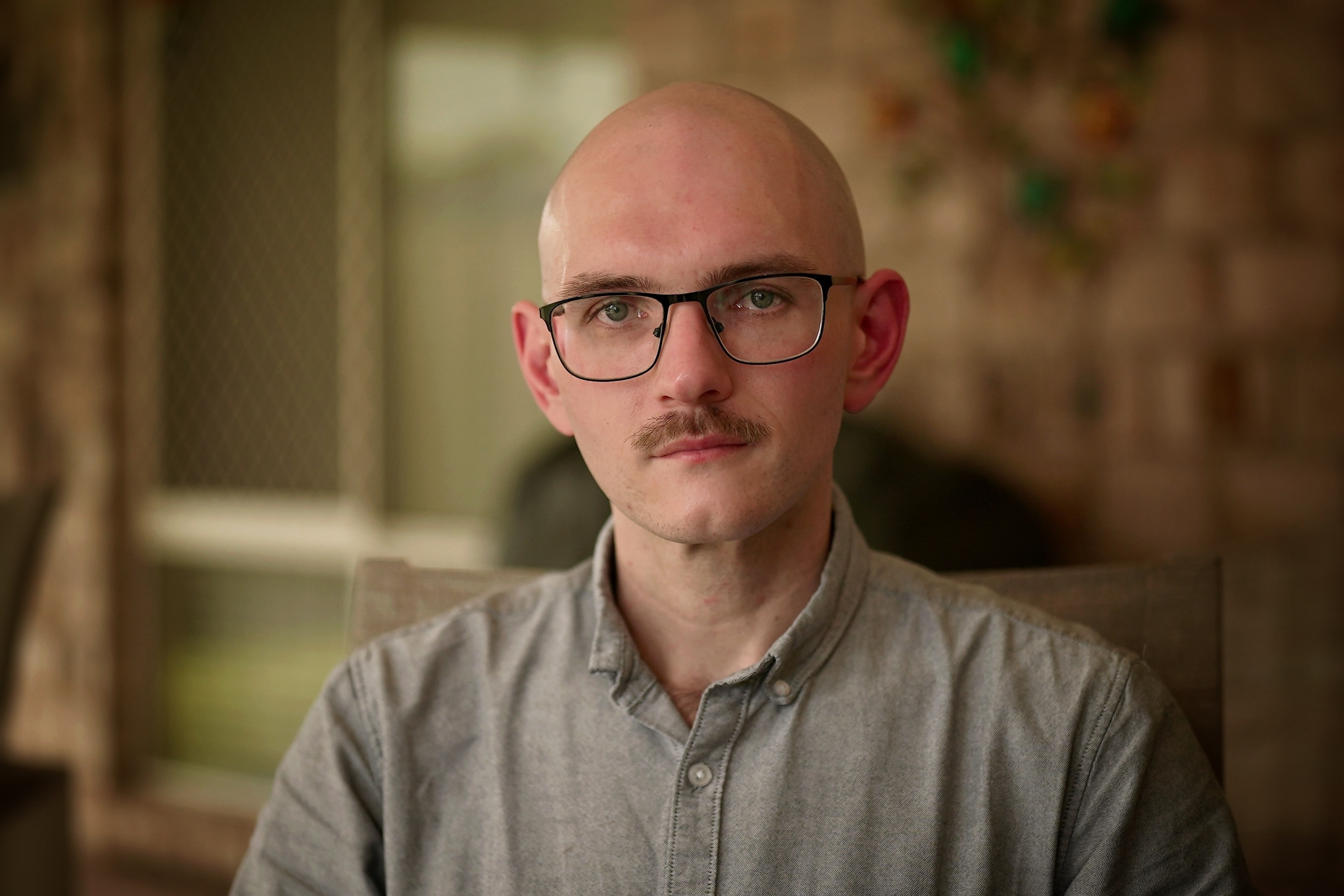 A man wearing glasses and a grey polo shirt looks towards the camera with a serious expression while sitting outside.