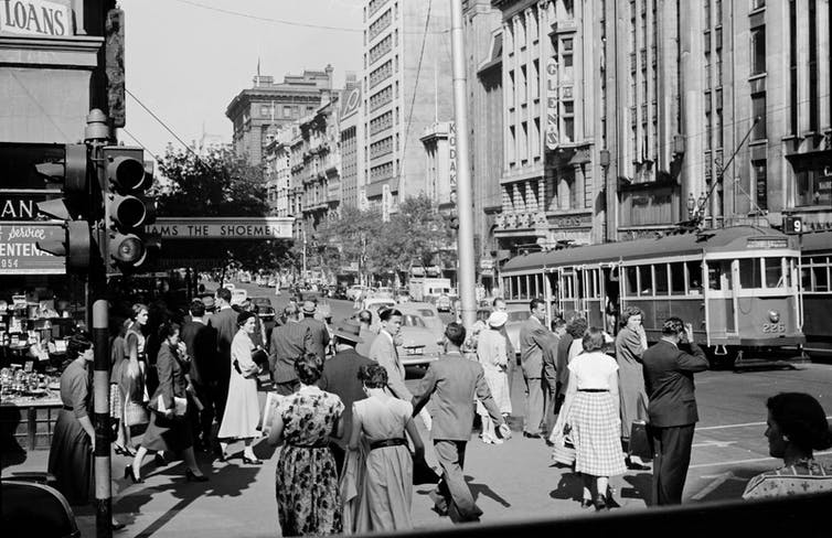Men in suits and women in dresses walk along Collins Street in a black and white image