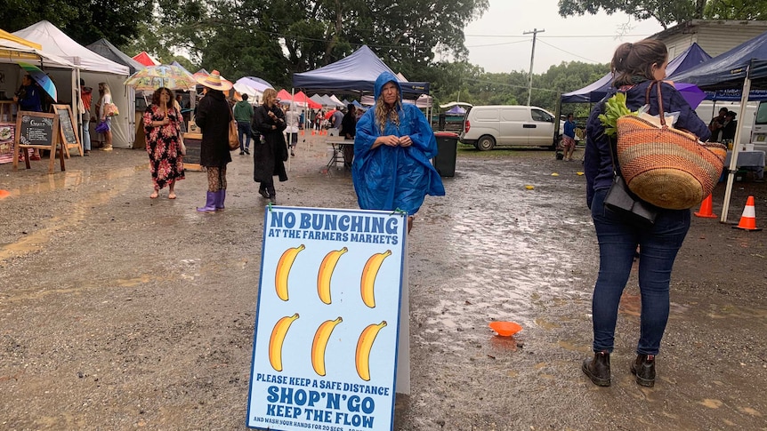 A sign reading "No Bunching at the Farmers Markets" stands in the mud at an open air bazaar in a regional area.