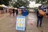 A sign reading "No Bunching at the Farmers Markets" stands in the mud at an open air bazaar in a regional area.