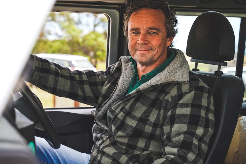 A man in a large fleece jacket smiles from the drivers seat of an old SUV.