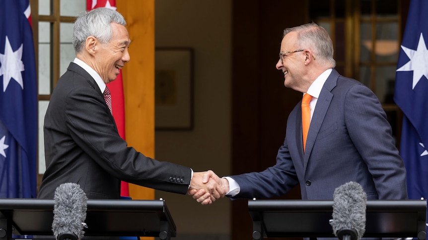 Two men in suits shake hands while standing in front of Australian flags.