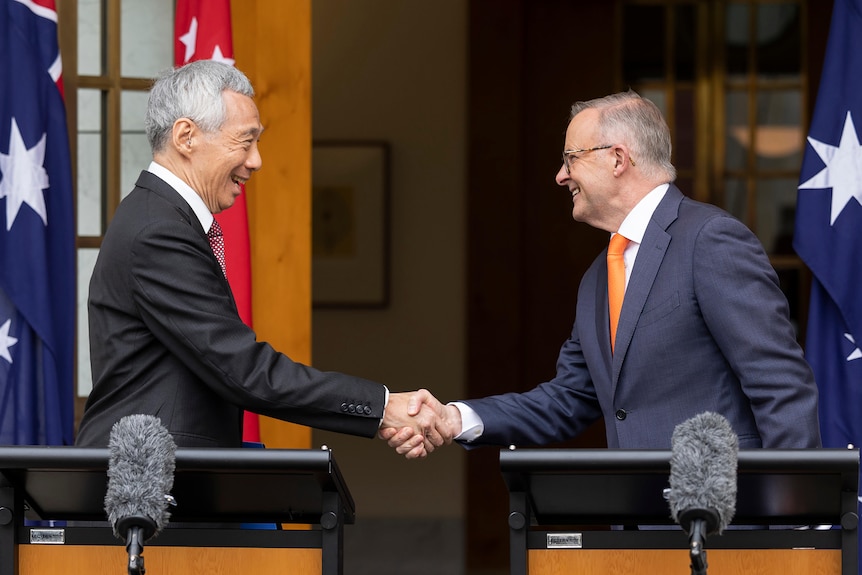 Two men in suits shake hands while standing in front of Australian flags.