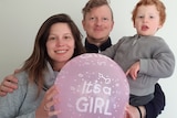 A woman with brown hair holds a balloon that reads "It's a girl" next to a man with red hair holding a toddler on his hip.