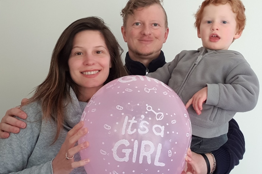 A woman with brown hair holds a balloon that reads "It's a girl" next to a man with red hair holding a toddler on his hip.