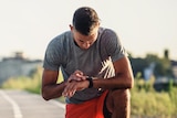 A young man on a road looks down at his watch. He is wearing workout gear. One knee is on the ground.