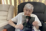 An elderly woman with grey hair looks at the camera. She is holding a picture of a soldier.