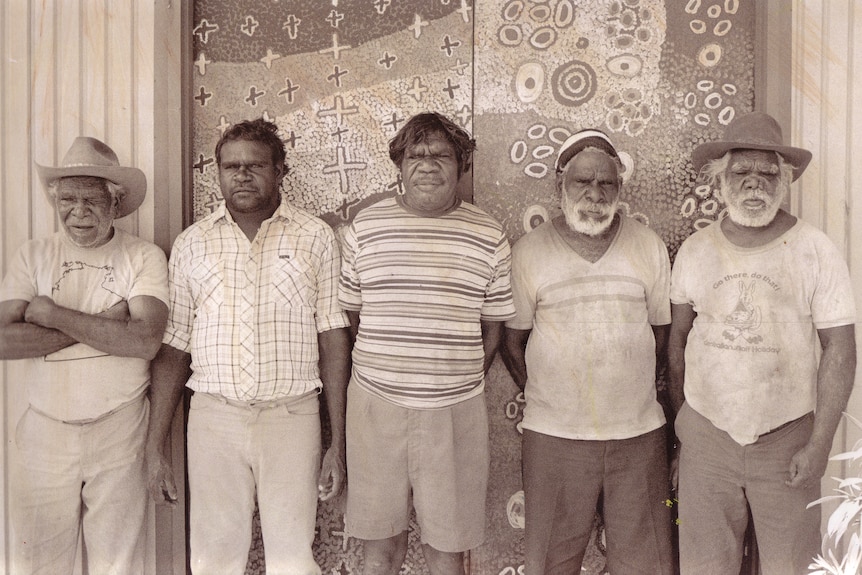 Black and white photo of five Warlpiri elders standing in front of classroom doors they have painted