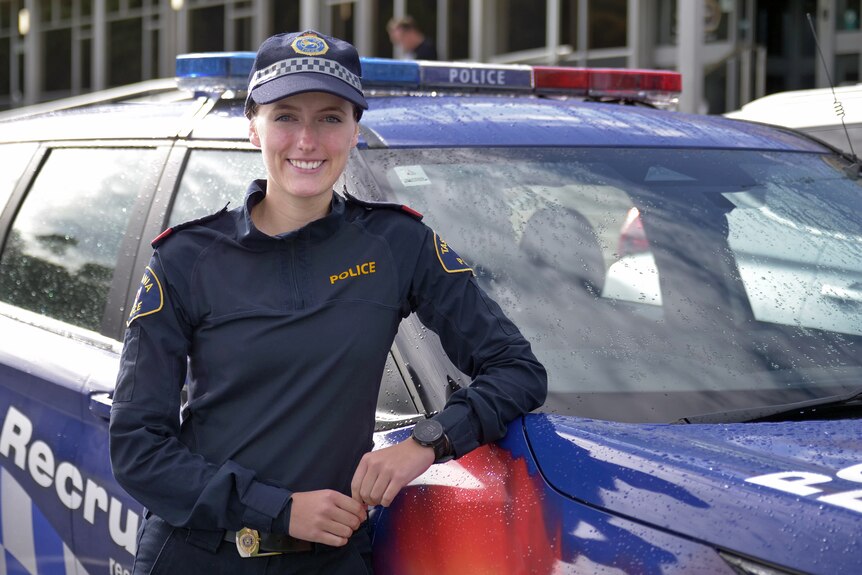 Female police recruit in navy uniform with her left arm leaning on a police vehicle.