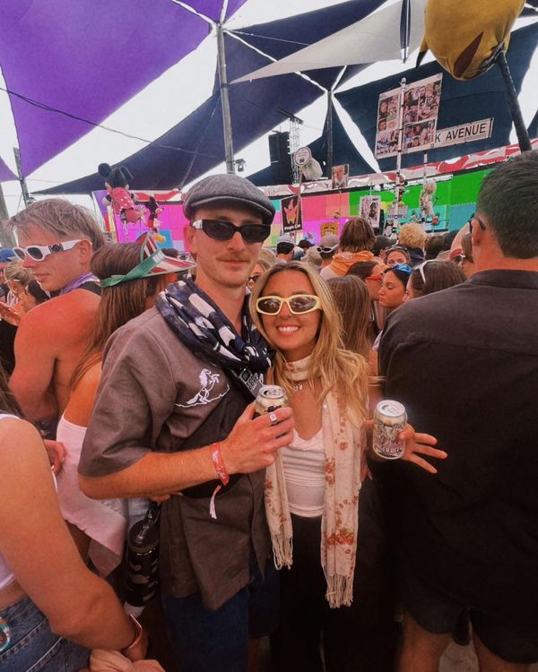 A man and a woman wearing sunglasses smile at the camera. The are in a crowd with multi-colour sails above them. 