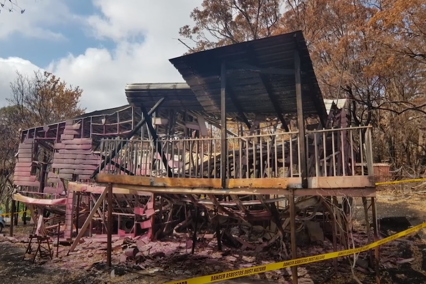 Shell of a house destroyed by fire with the roof collapsed and tape in front saying Danger Asbestos Dust Hazard