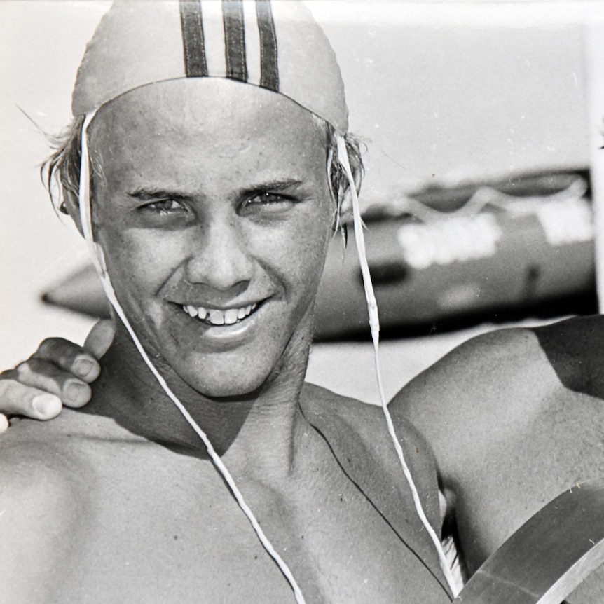 A black-and-white photo of a young man in surf livesaving cap is embraced by his father.