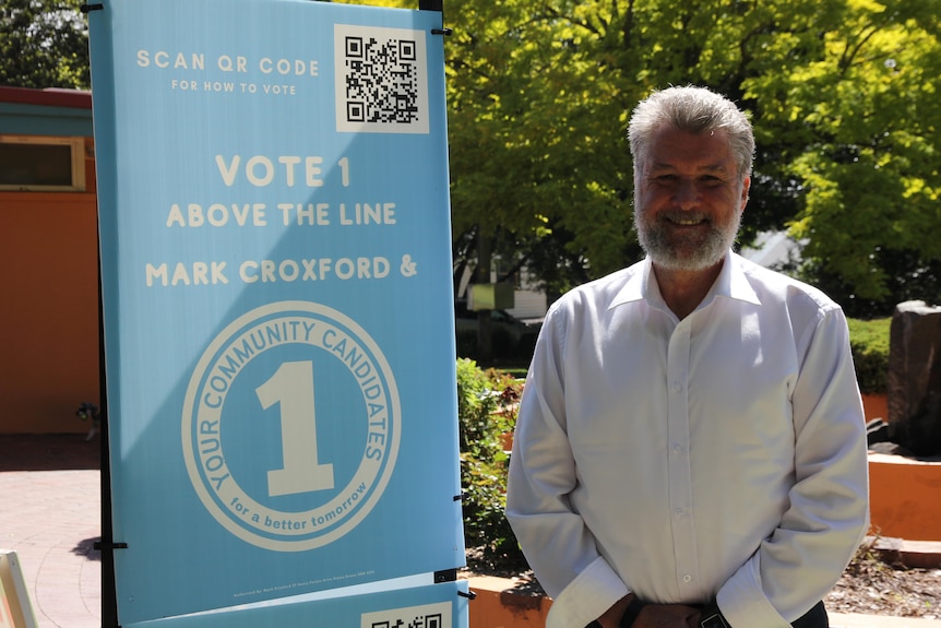 An older man in a business shirt stands next to an election banner.