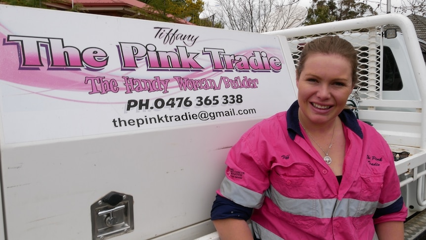 A young woman wearing a pink high-visibility shirt stands in front of a work ute labelled with the logo The Pink Tradie.