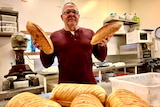 A man stands in a bakery holding up two loaves of bread.
