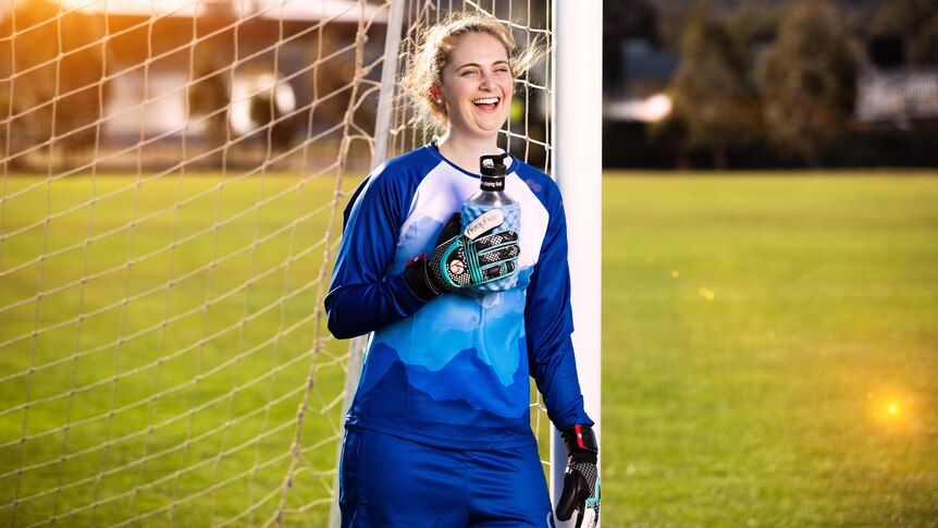 Female soccer player leaning against the right goal post smiling while  wearing her women's specific goalie gloves 