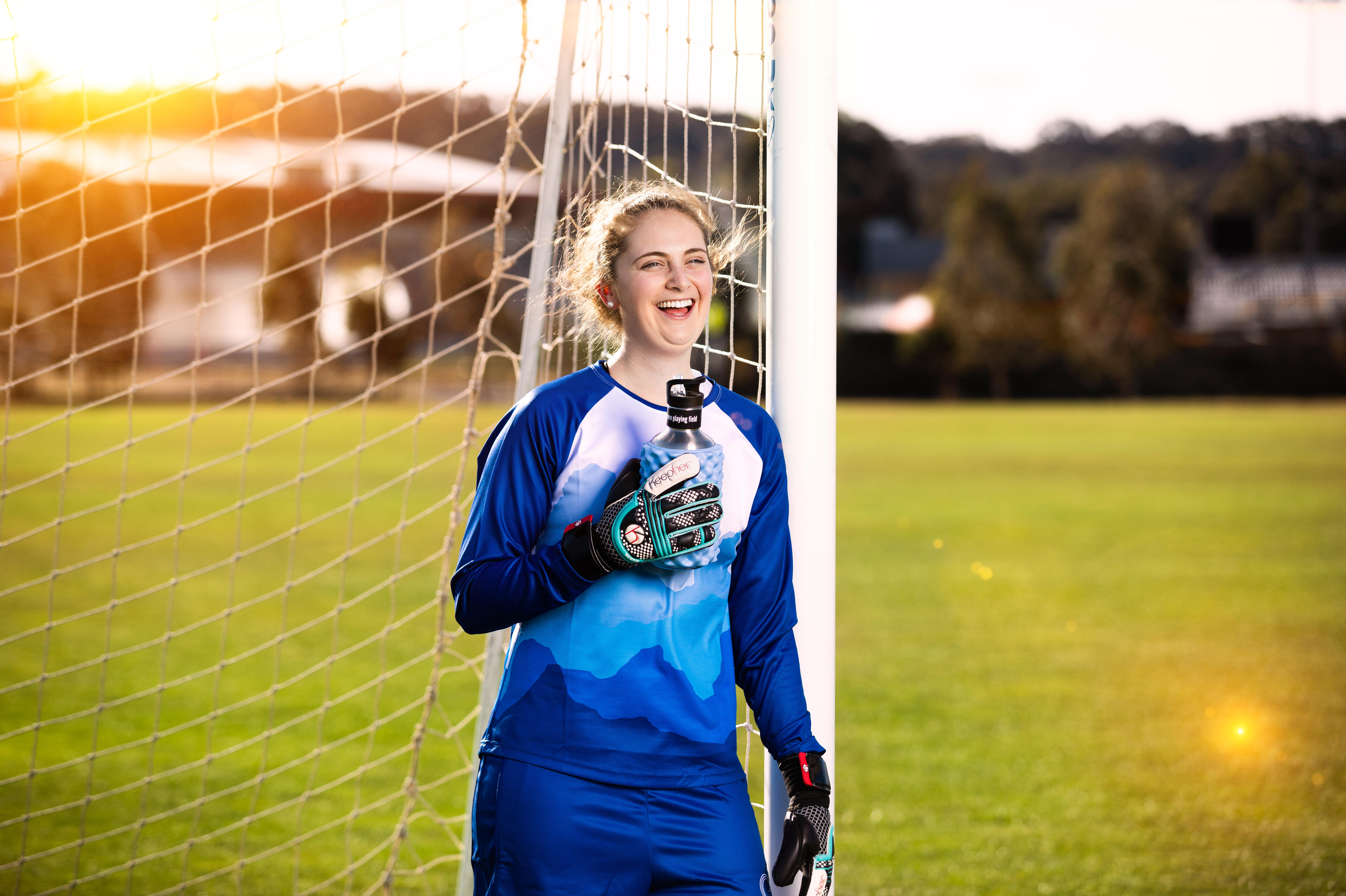 Female soccer player leaning against the right goal post smiling while  wearing her women's specific goalie gloves 