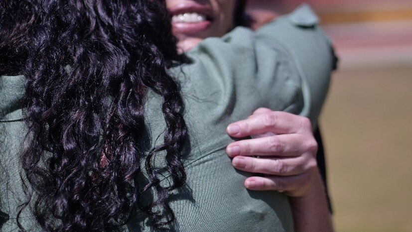 A woman with black curly hair faces away from the camera as she hugs another woman, outdoors in the midday sun. 