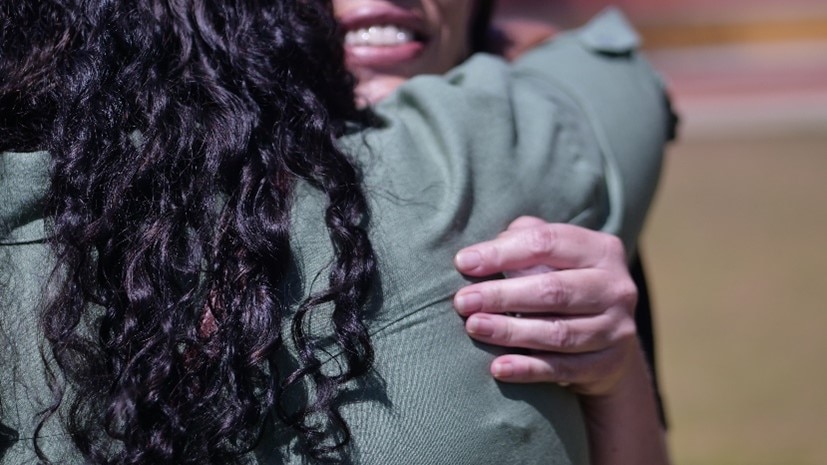 A woman with black curly hair faces away from the camera as she hugs another woman, outdoors in the midday sun. 