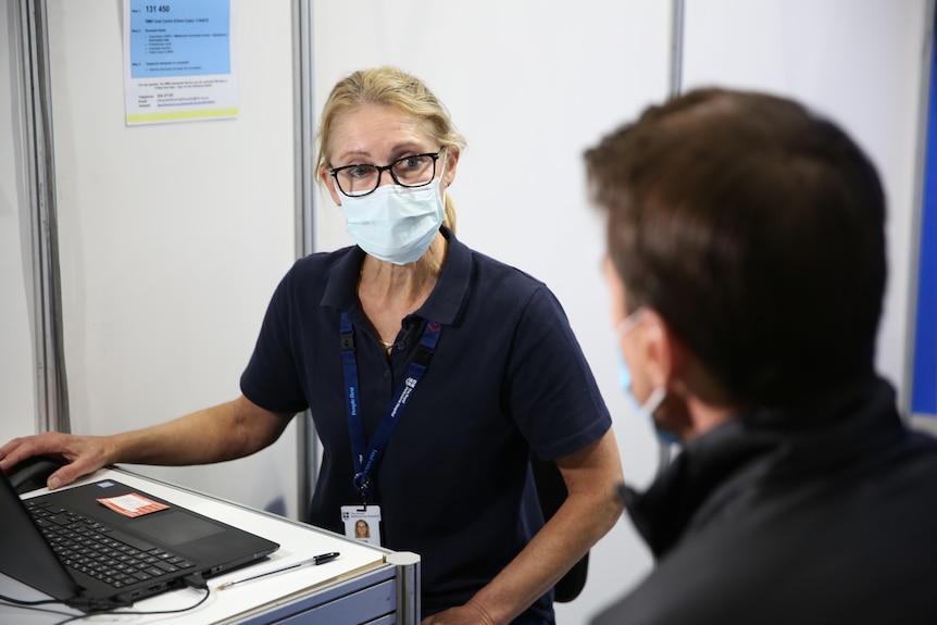 A vaccine nurse wearing a mask speaks to an out-of-focus Michael Rowland.