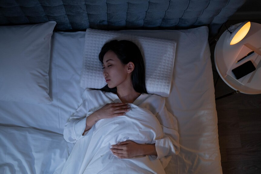 A woman lies in bed in the dark, with a bedside lamp illuminating a notebook.