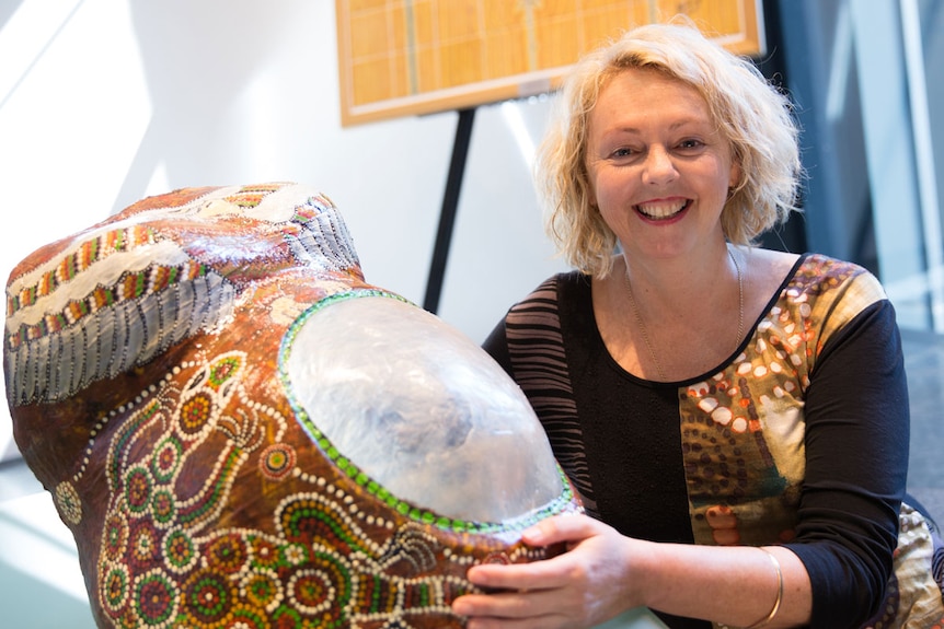 Dr Kym Rae smiles and holds a colourfully painted plaster cast of an Indigenous woman's pregnant belly.