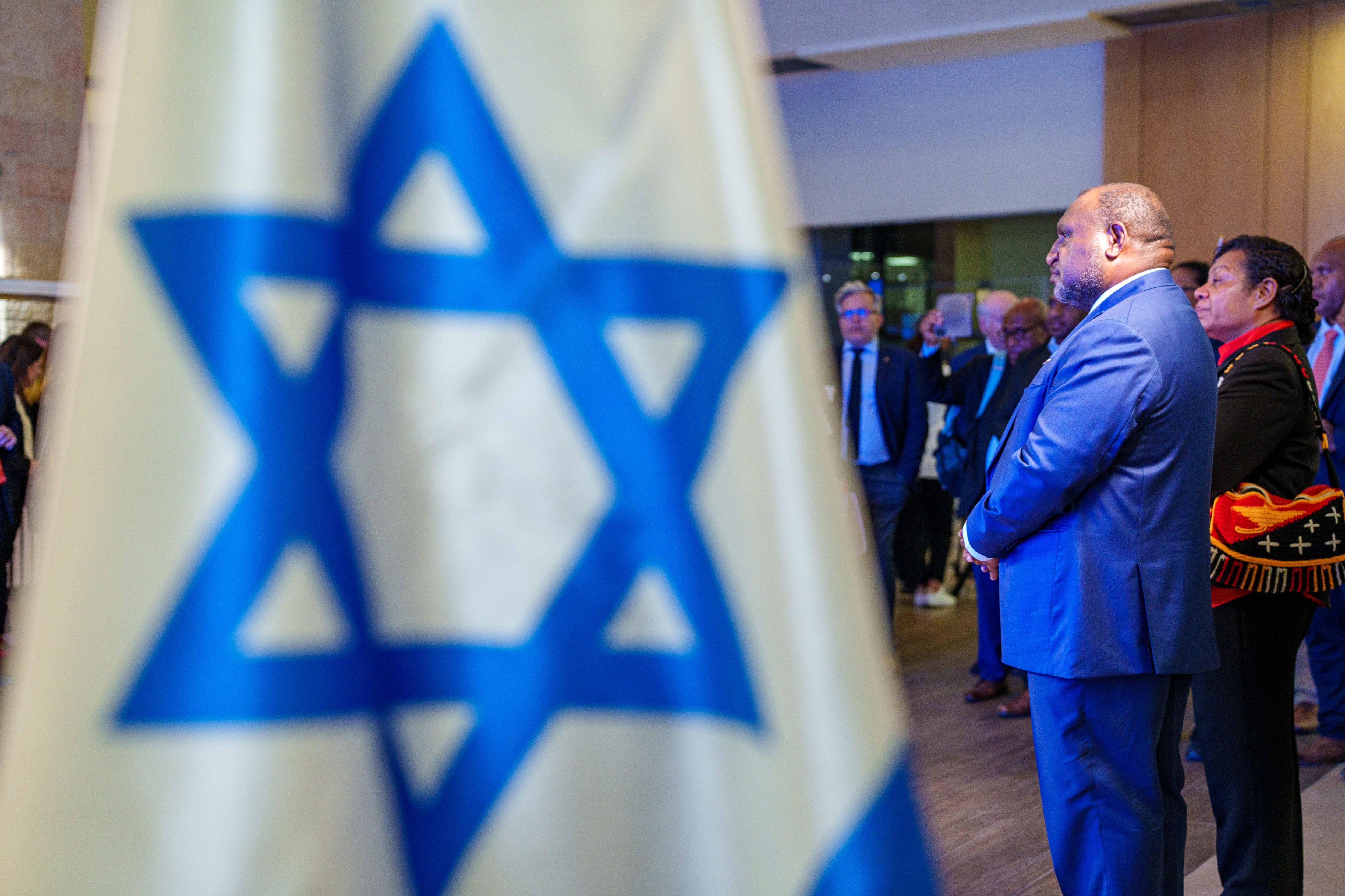 Man in blue suit stands in front of Israeli flag 