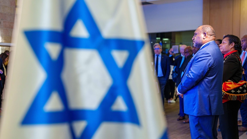 Man in blue suit stands in front of Israeli flag 